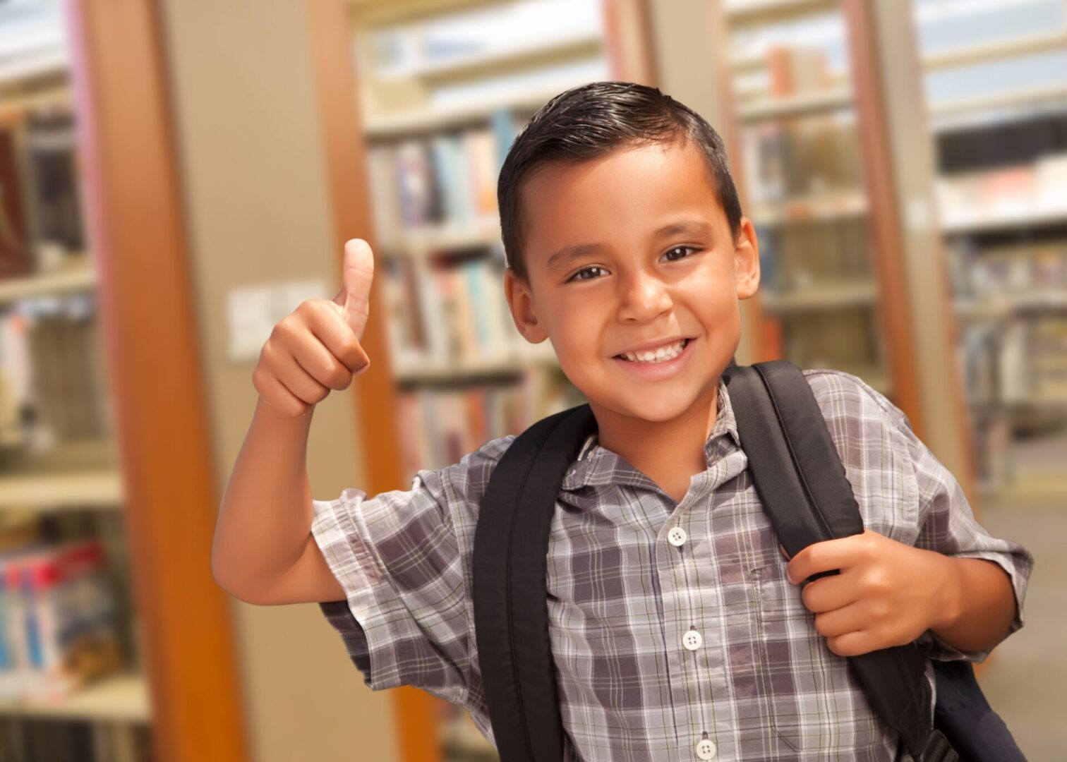 A boy with backpack giving thumbs up in library.